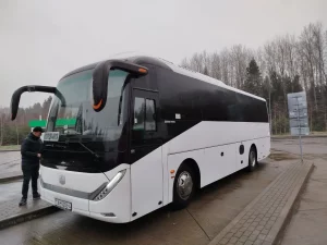 Вялікі белы аўтобус Zhong Tong, прыпаркаваны на мокрым тратуары каля дрэў, з чалавекам, які стаіць побач, адкідвае адзінокую постаць на фоне хмарнага неба трасы Гродна-Мінск.